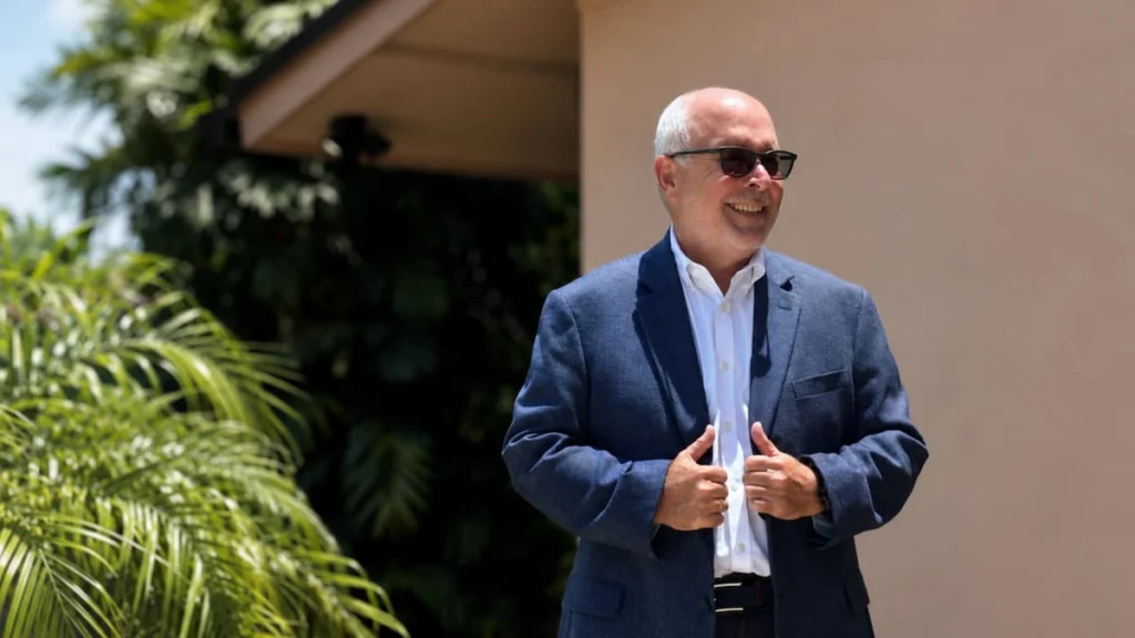 Rafale de Leon con traje azul y camisa blanca sonriendo frente a una vivienda y vegetación tropical