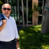 Hombre con chaqueta azul posando al aire libre en un entorno residencial rodeado de vegetación tropical