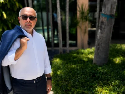 Hombre con chaqueta azul posando al aire libre en un entorno residencial rodeado de vegetación tropical