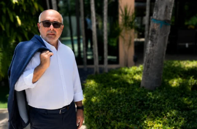 Hombre con chaqueta azul posando al aire libre en un entorno residencial rodeado de vegetación tropical