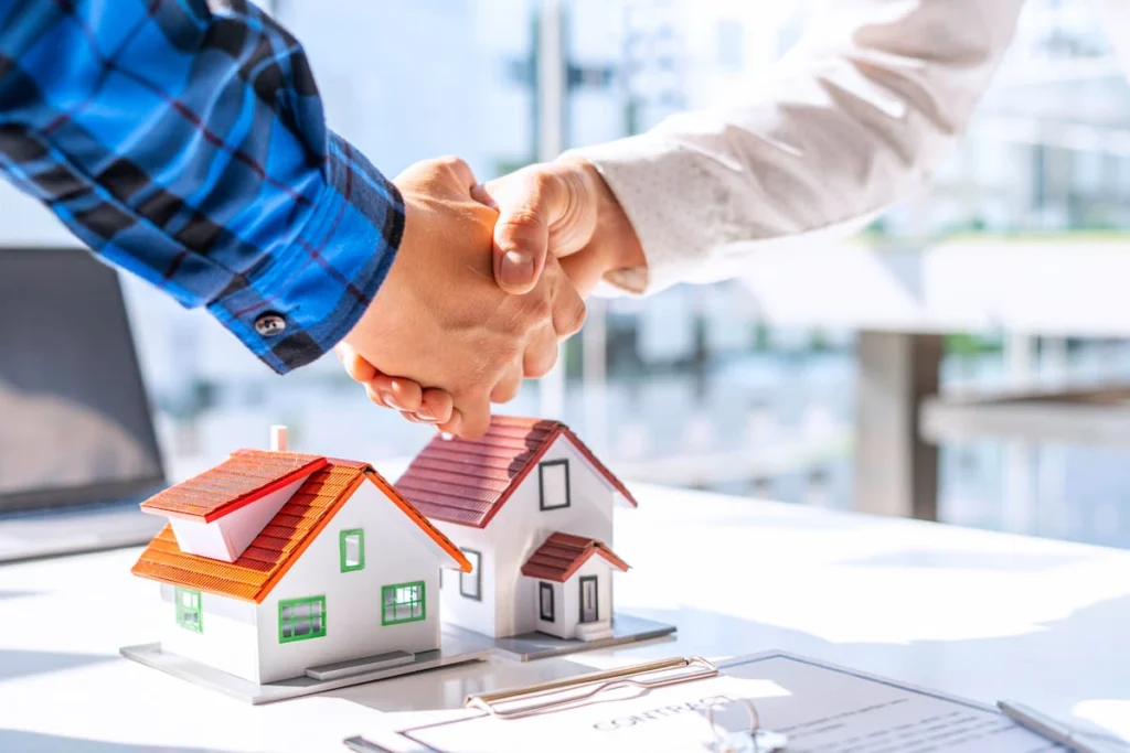 Dos personas estrechan la mano sobre una mesa con maquetas de viviendas y un contrato inmobiliario