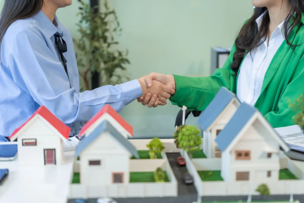 Dos personas estrechan la mano frente a varias maquetas de casas como símbolo de acuerdo o negociación inmobiliaria