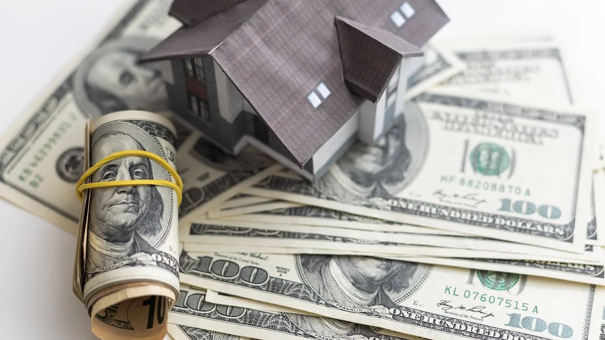 Maqueta de una casa sobre billetes de cien dólares junto a un rollo de dinero como símbolo de financiamiento o inversión inmobiliaria