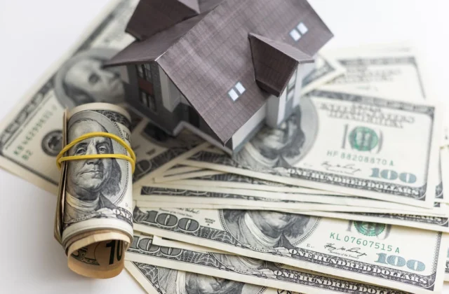 Maqueta de una casa sobre billetes de cien dólares junto a un rollo de dinero como símbolo de financiamiento o inversión inmobiliaria
