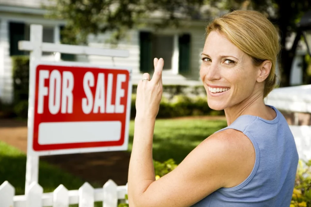 Mujer sonriente frente a una vivienda con un cartel de “For Sale”, como símbolo de una propiedad puesta a la venta