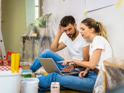 Pareja revisando el presupuesto de su remodelación en una laptop, sentados en el suelo entre cubetas de pintura