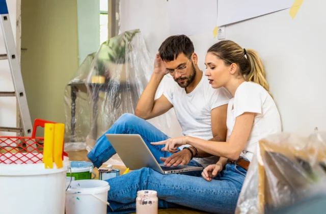 Pareja revisando el presupuesto de su remodelación en una laptop, sentados en el suelo entre cubetas de pintura
