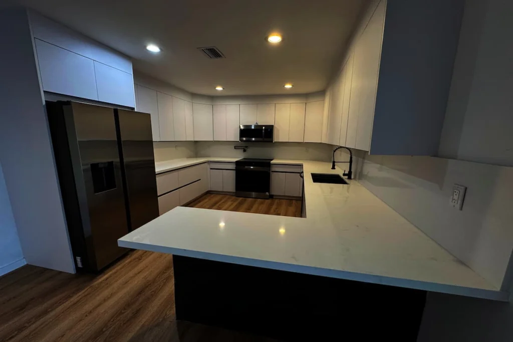 Cocina renovada con gabinetes blancos, encimeras claras, electrodomésticos oscuros e iluminación empotrada
