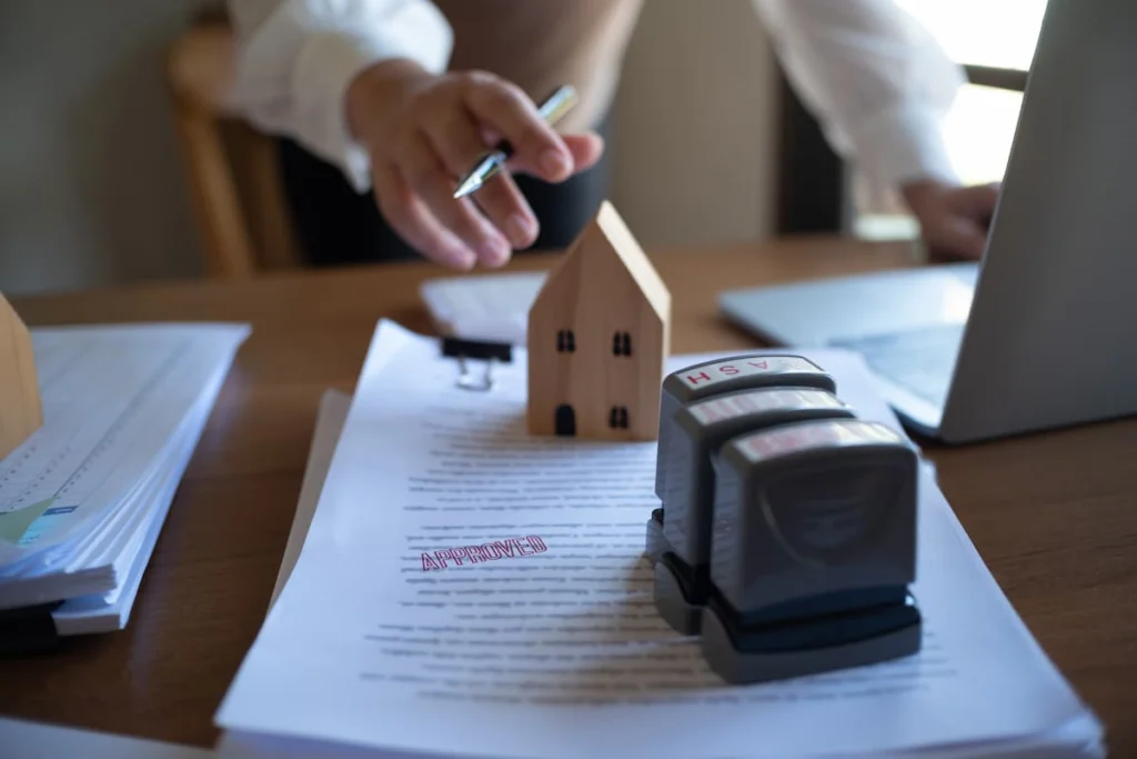 Contrato de compra de vivienda con sello de aprobado junto a una maqueta de casa y una computadora portátil