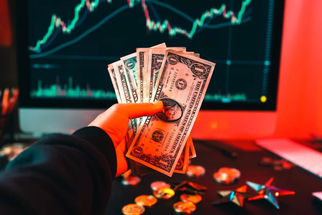 Mano sosteniendo varios billetes de dólar frente a una pantalla con gráficos de cotizaciones financieras