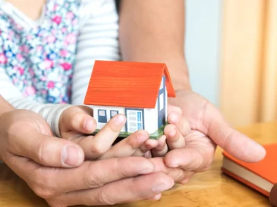 Manos de un adulto y un niño sosteniendo juntos una pequeña casa, como símbolo de vivienda, familia y patrimonio