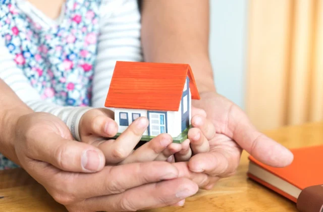 Manos de un adulto y un niño sosteniendo juntos una pequeña casa, como símbolo de vivienda, familia y patrimonio