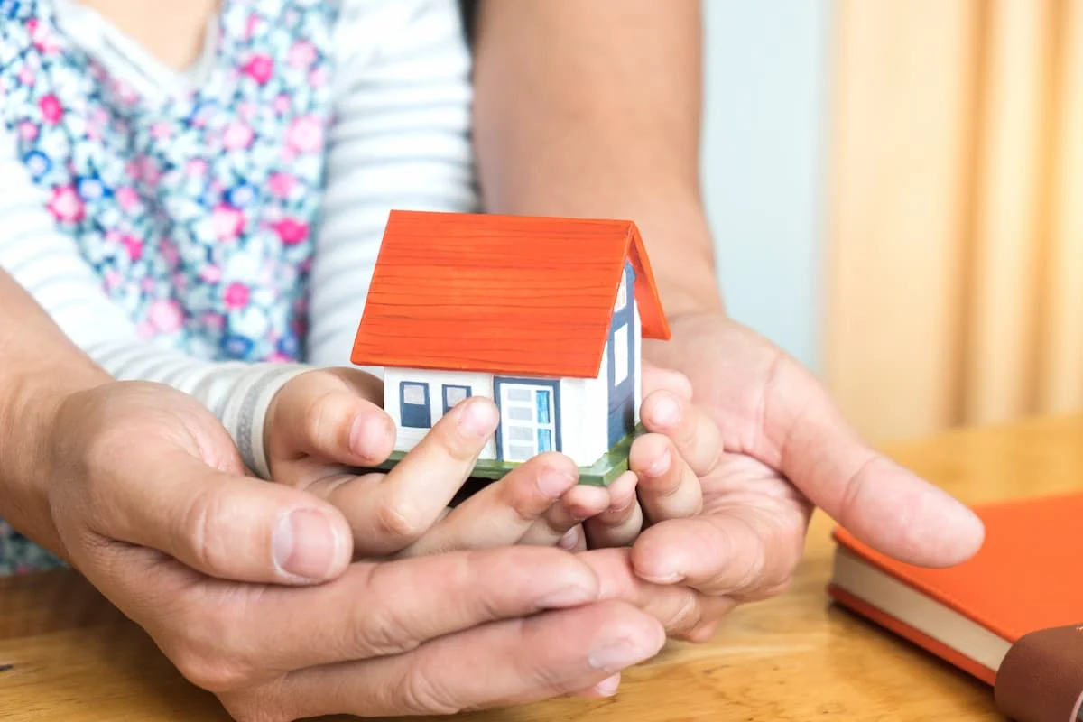 Manos de un adulto y un niño sosteniendo juntos una pequeña casa, como símbolo de vivienda, familia y patrimonio