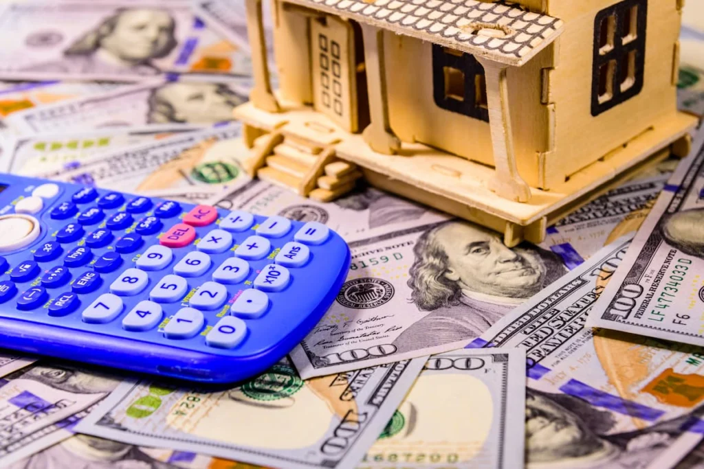 Maqueta de una vivienda junto a una calculadora y billetes de cien dólares como símbolo de costos y finanzas del hogar