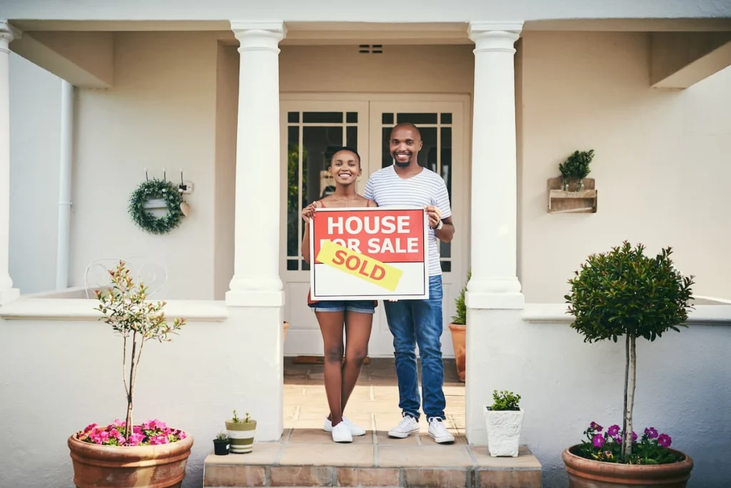 Pareja sonriente frente a su nueva vivienda sosteniendo un cartel de propiedad vendida