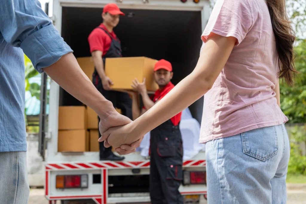 Una pareja tomada de la mano observa a dos trabajadores que descargan cajas de cartón de un camión de mudanzas estacionado frente a una vivienda.