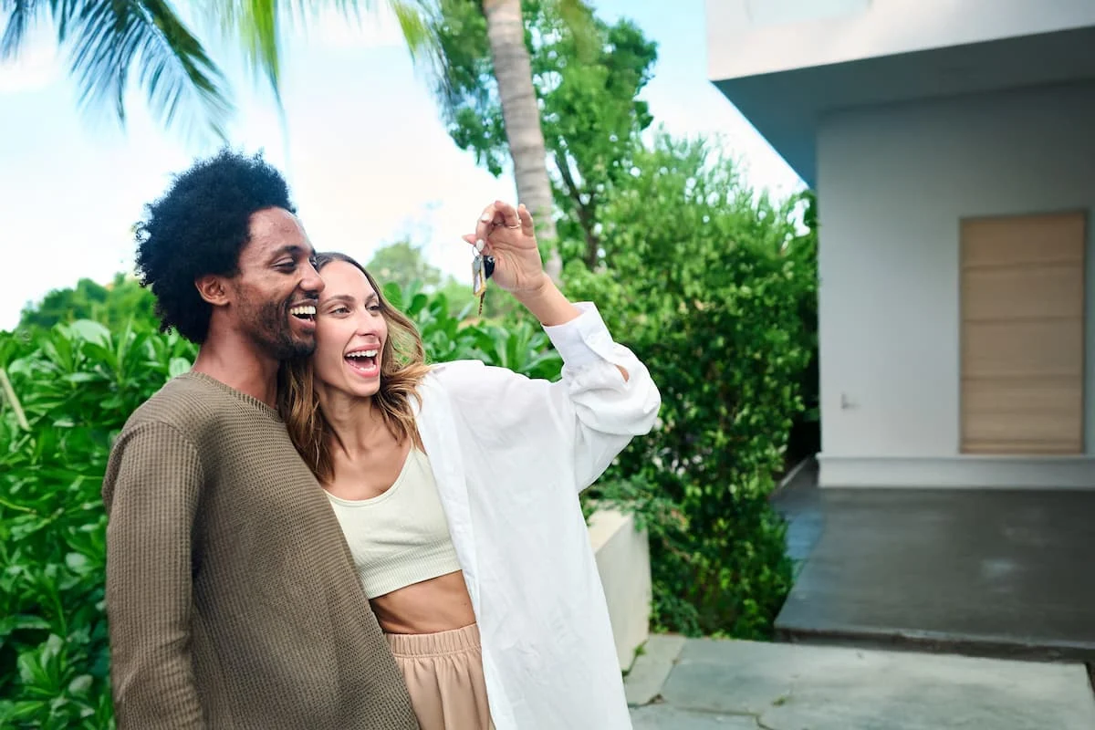 Pareja sonriente mostrando las llaves de su nueva casa frente a la entrada de una vivienda