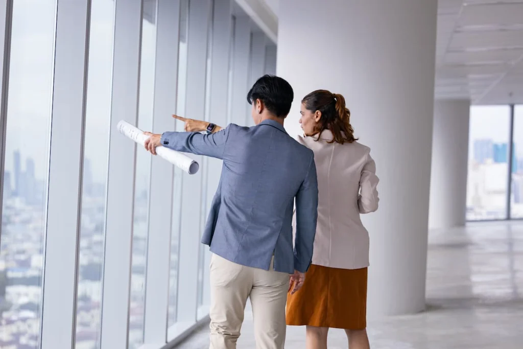 Dos profesionales revisando planos y señalando los ventanales de un espacio comercial moderno con vistas a la ciudad