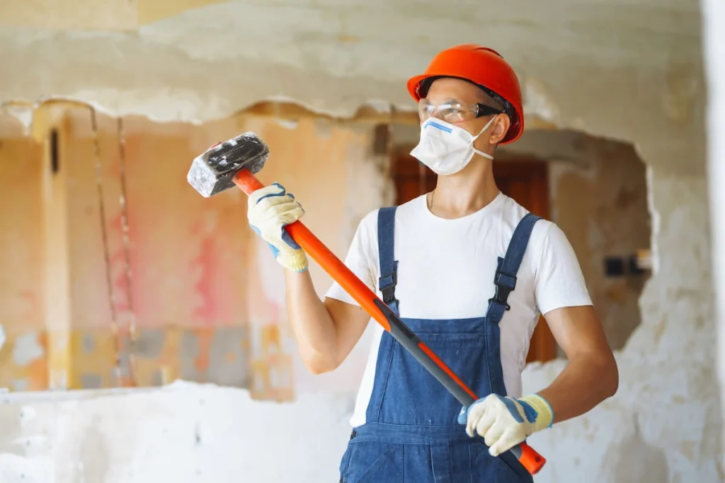 Trabajador con casco naranja, gafas de protección, mascarilla y guantes sostiene un mazo de demolición en el interior de una vivienda con paredes parcialmente descubiertas.