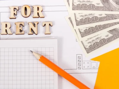 Letras de madera con el texto “FOR RENT” junto a billetes de cien dólares, un lápiz y documentos sobre una mesa.