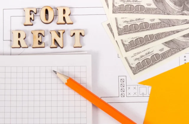 Letras de madera con el texto “FOR RENT” junto a billetes de cien dólares, un lápiz y documentos sobre una mesa.
