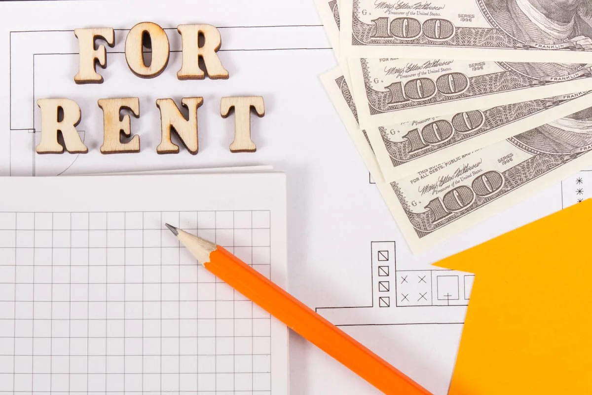 Letras de madera con el texto “FOR RENT” junto a billetes de cien dólares, un lápiz y documentos sobre una mesa.