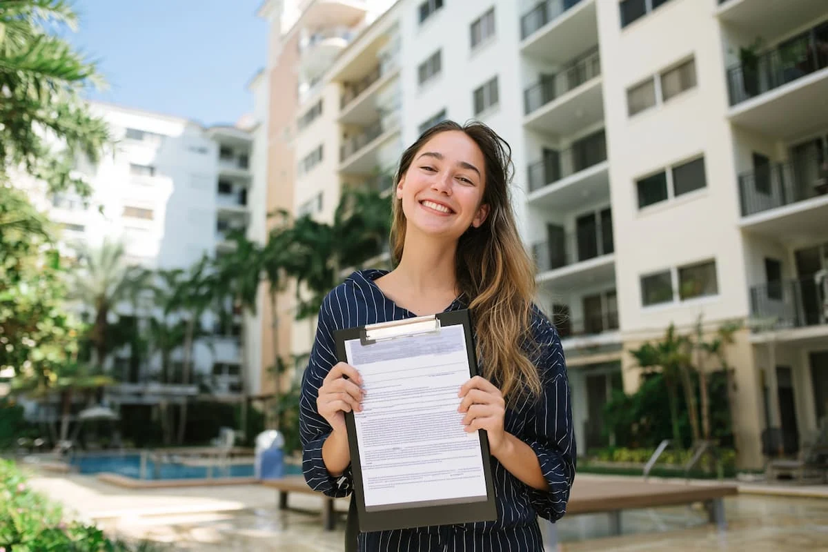 Mujer sostiene una carpeta con documentos frente a un edificio residencial con balcones, piscina y áreas verdes.