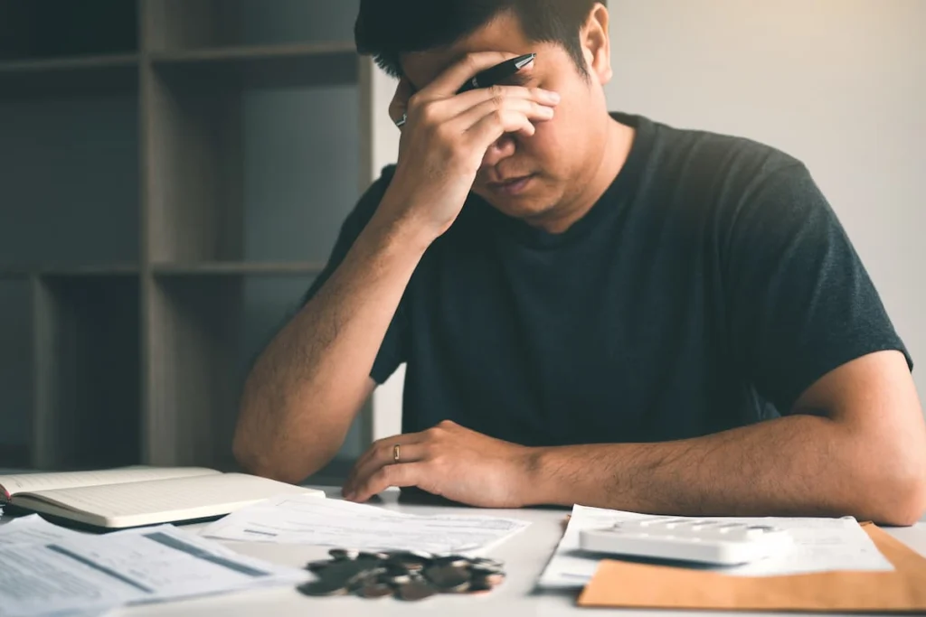 Hombre sentado frente a documentos, una calculadora y monedas se cubre el rostro con una mano mientras revisa sus finanzas.