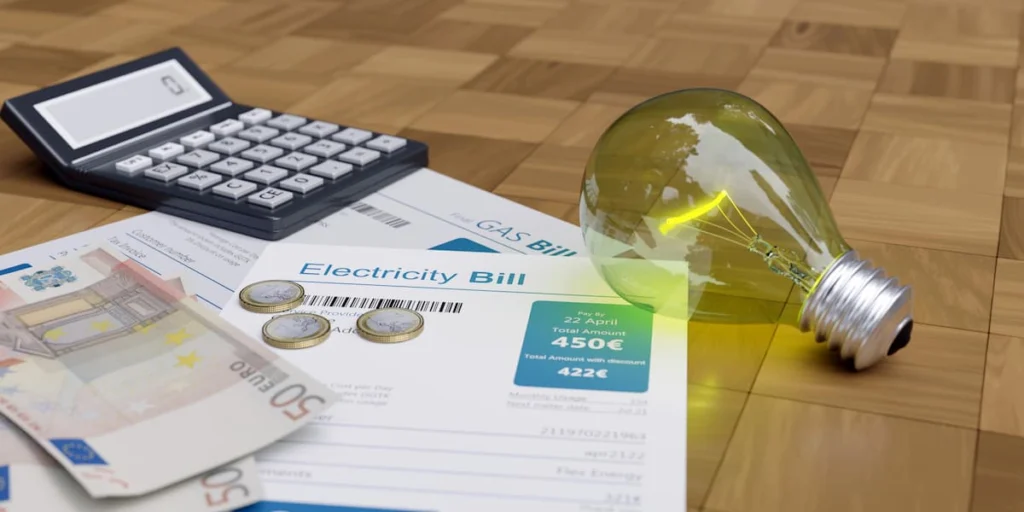 Factura de electricidad junto a una calculadora, monedas, billetes y una bombilla sobre una mesa.