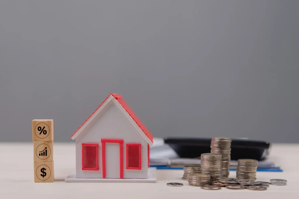 Maqueta de una vivienda junto a pilas de monedas y bloques con símbolos de porcentaje, crecimiento y dólar, en referencia al financiamiento inmobiliario.