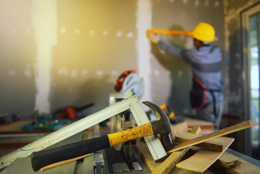 Martillo, escuadra, piezas de madera y otras herramientas descansan sobre una sierra de mesa mientras un trabajador realiza mediciones en una pared en remodelación.