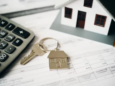 Calculadora, llave y figuras de casas colocadas sobre documentos financieros relacionados con una vivienda.