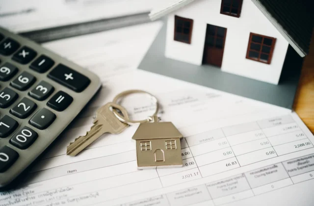 Calculadora, llave y figuras de casas colocadas sobre documentos financieros relacionados con una vivienda.