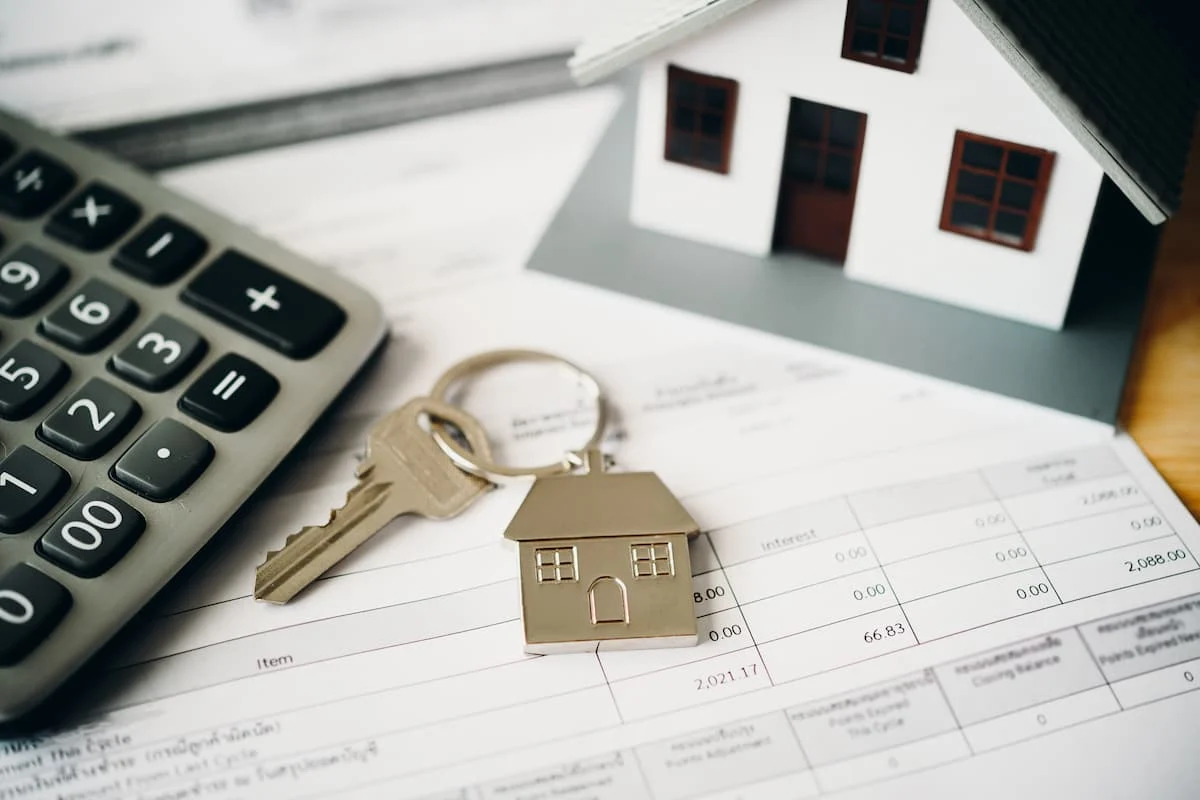 Calculadora, llave y figuras de casas colocadas sobre documentos financieros relacionados con una vivienda.