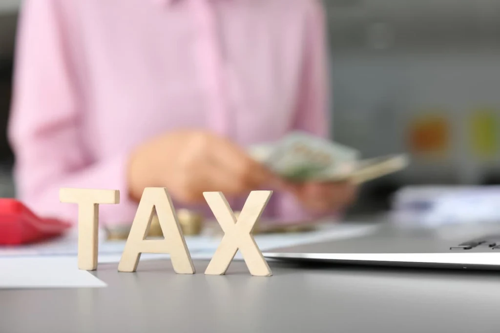Letras de madera con la palabra “TAX” en primer plano mientras una persona cuenta dinero junto a documentos y una computadora portátil.