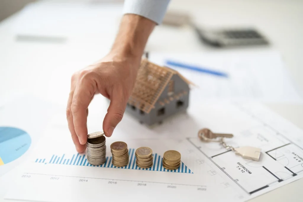 Mano coloca monedas apiladas sobre un gráfico financiero junto a una maqueta de vivienda, unas llaves y planos.