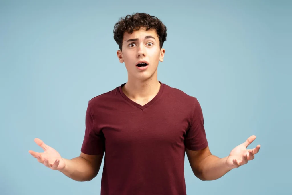 Joven con expresión de sorpresa mantiene los brazos abiertos y las palmas hacia arriba sobre un fondo azul claro.