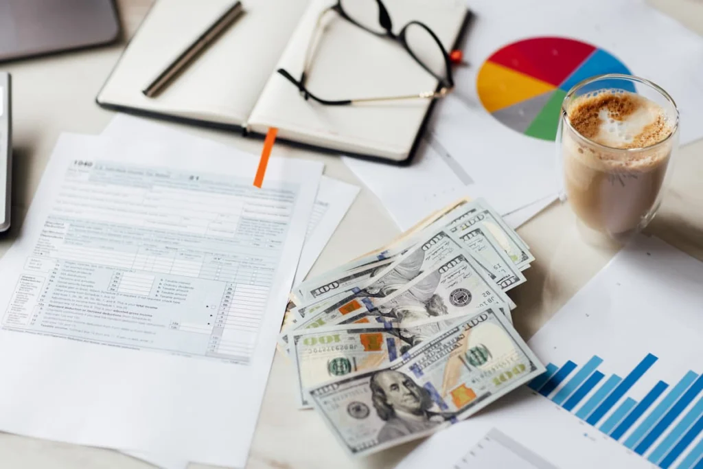 Billetes de cien dólares sobre documentos financieros con gráficos, formularios, una libreta, gafas y una taza de café.
