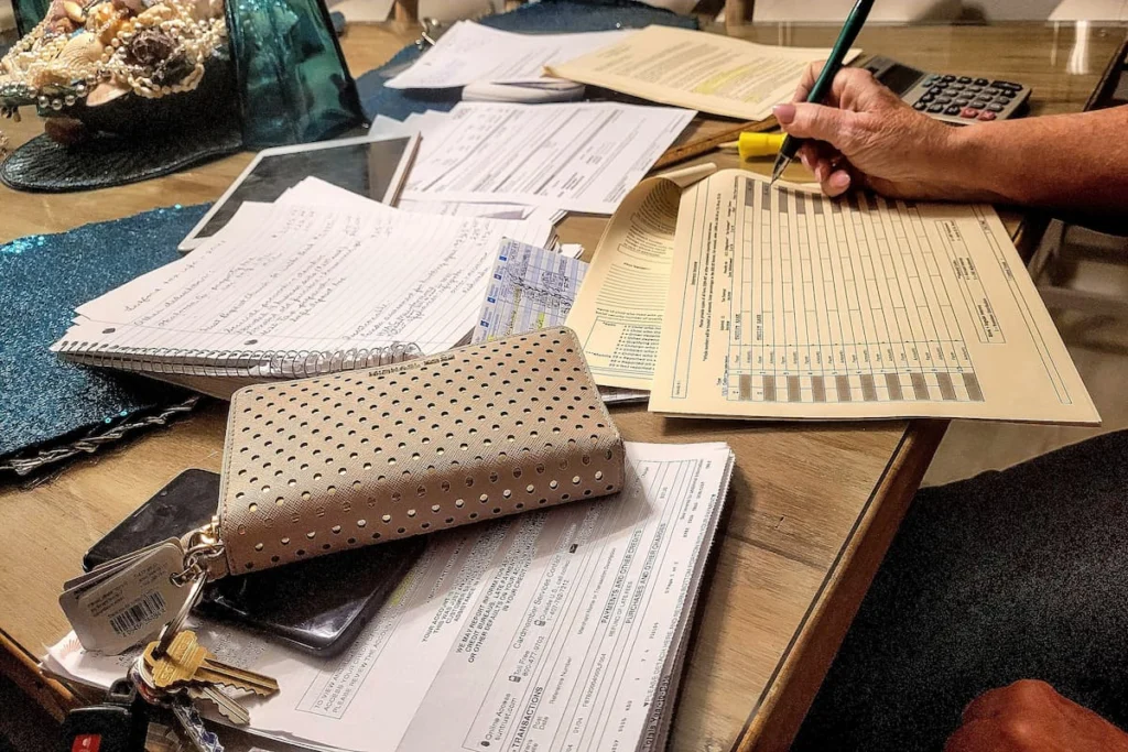 Persona completa formularios y revisa documentos financieros sobre una mesa con una calculadora, cuaderno, llaves y papeles.