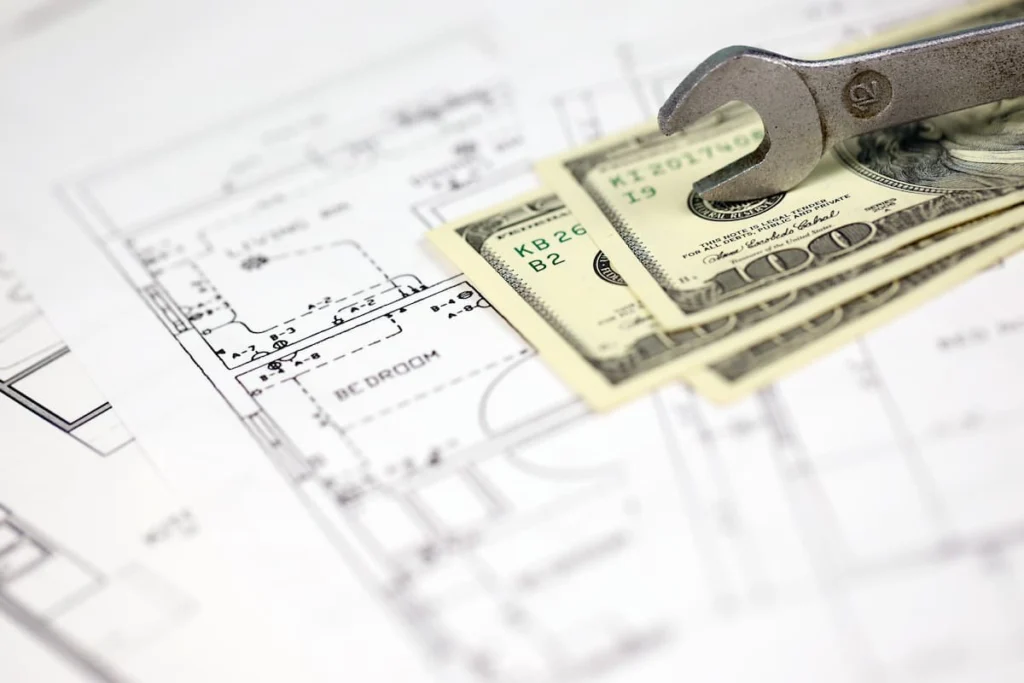 Billetes de cien dólares y una llave inglesa sobre planos de construcción, en referencia a los costos y al presupuesto de una obra.