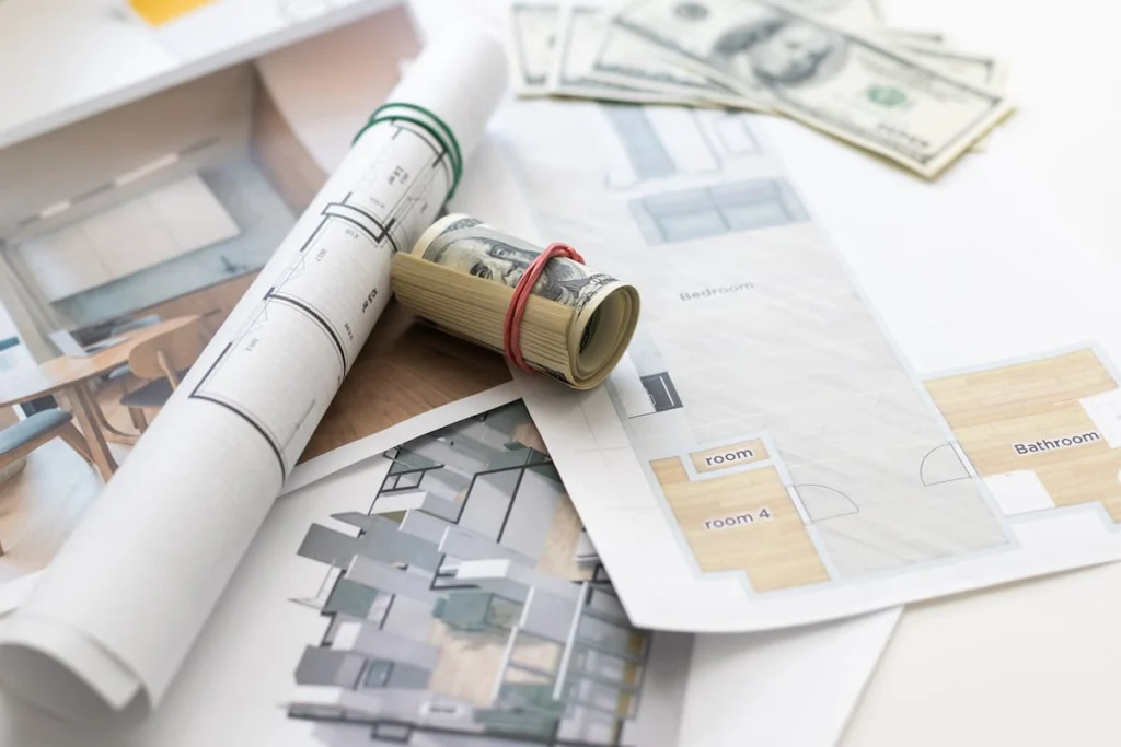 Planos de una vivienda junto a billetes de cien dólares y un rollo de dinero, en referencia al presupuesto de una remodelación.
