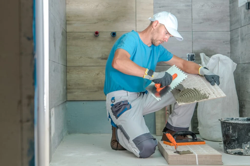 Trabajador aplica mortero adhesivo sobre una baldosa durante la instalación de revestimientos en un baño en remodelación.