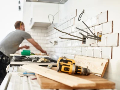 Trabajador realiza trabajos de remodelación en una cocina con azulejos parcialmente instalados, cables eléctricos expuestos y herramientas sobre la encimera.