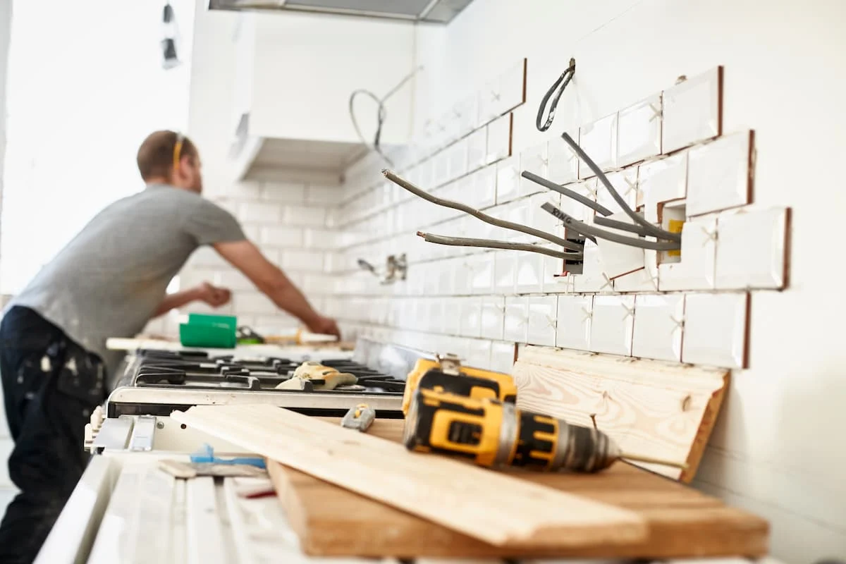Trabajador realiza trabajos de remodelación en una cocina con azulejos parcialmente instalados, cables eléctricos expuestos y herramientas sobre la encimera.
