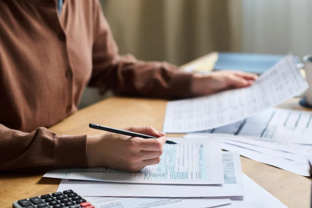 Persona revisa y completa formularios financieros sobre una mesa con varios documentos y una calculadora.