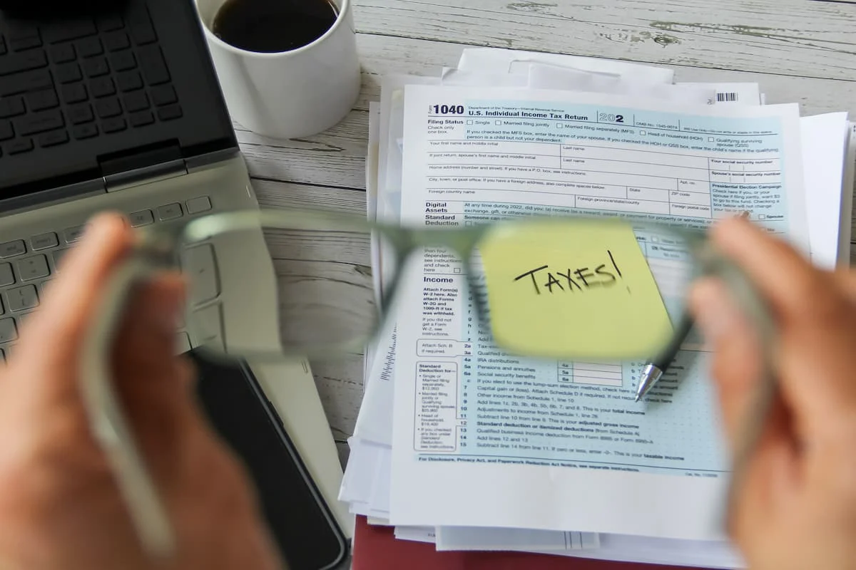 Persona revisa con una lupa un formulario de impuestos de Estados Unidos junto a una nota con la palabra “TAXES”, una computadora portátil y documentos.