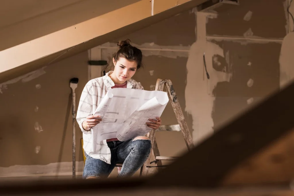Mujer revisa planos de remodelación junto a una escalera en el interior de una vivienda con paredes en proceso de acabado.