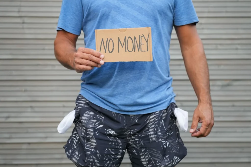 Persona con los bolsillos vacíos sostiene un cartel de cartón con el texto “NO MONEY”.