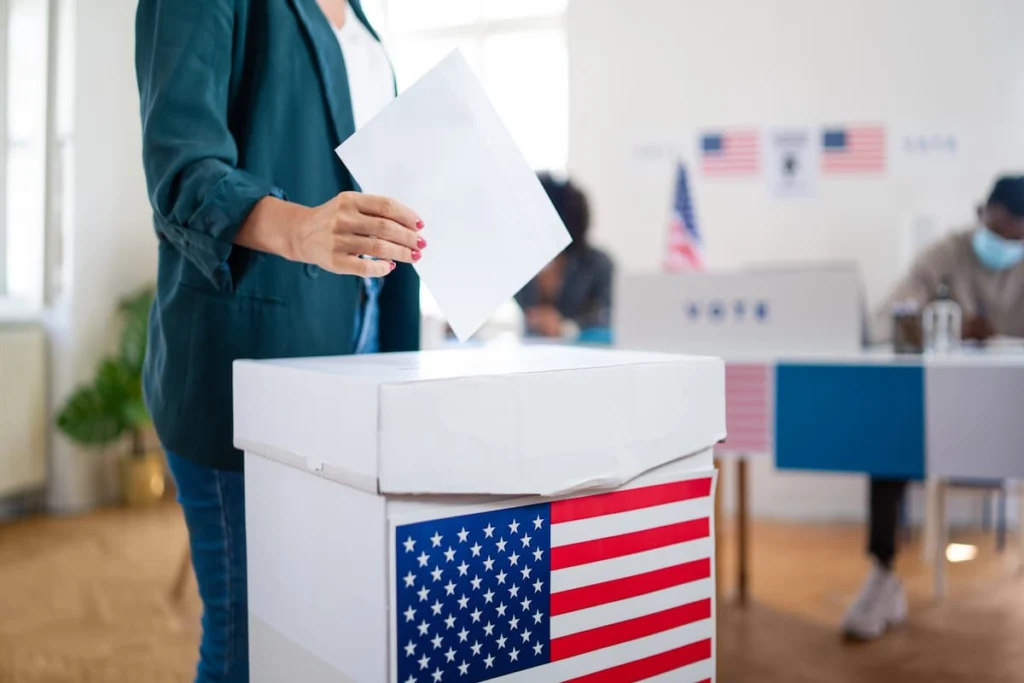 Persona deposita una papeleta en una urna decorada con la bandera de Estados Unidos dentro de un centro de votación. 